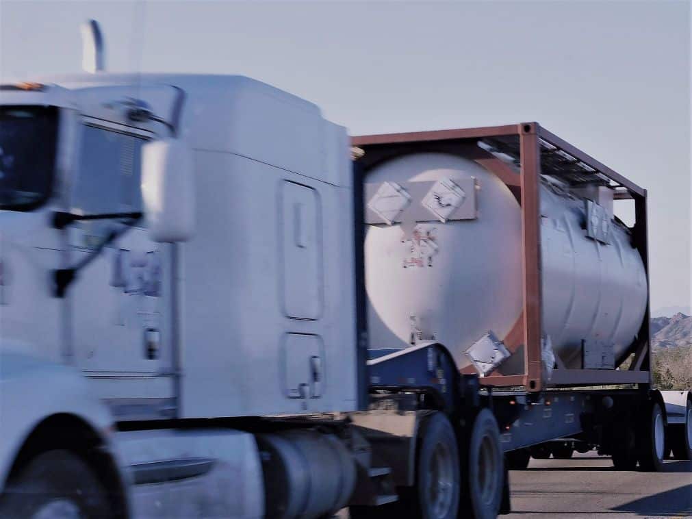 White semi-truck towing a large cylindrical tank marked with hazardous material signs on a highway.